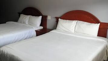 Ett hotellrum med två separata sängar, båda med vita sängkläder och sänggavlar i trä. Det finns två sängbord, det ena med en lampa och det andra med ett eluttag. Väggarna är målade i en neutral färg, och det finns en taklampa.