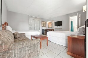 Une chambre d’hôtel avec un canapé, un lit, une table de chevet en bois, une télévision et une fenêtre avec des stores.