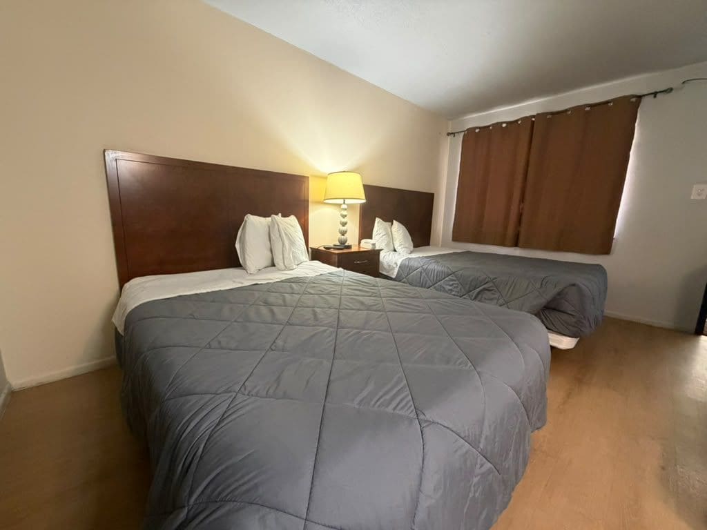 A hotel room with two beds, a nightstand, a lamp, and a window with curtains.