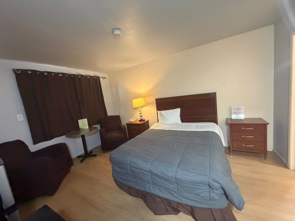 A hotel room with a large bed, a desk, a chair, a lamp, and a dresser.