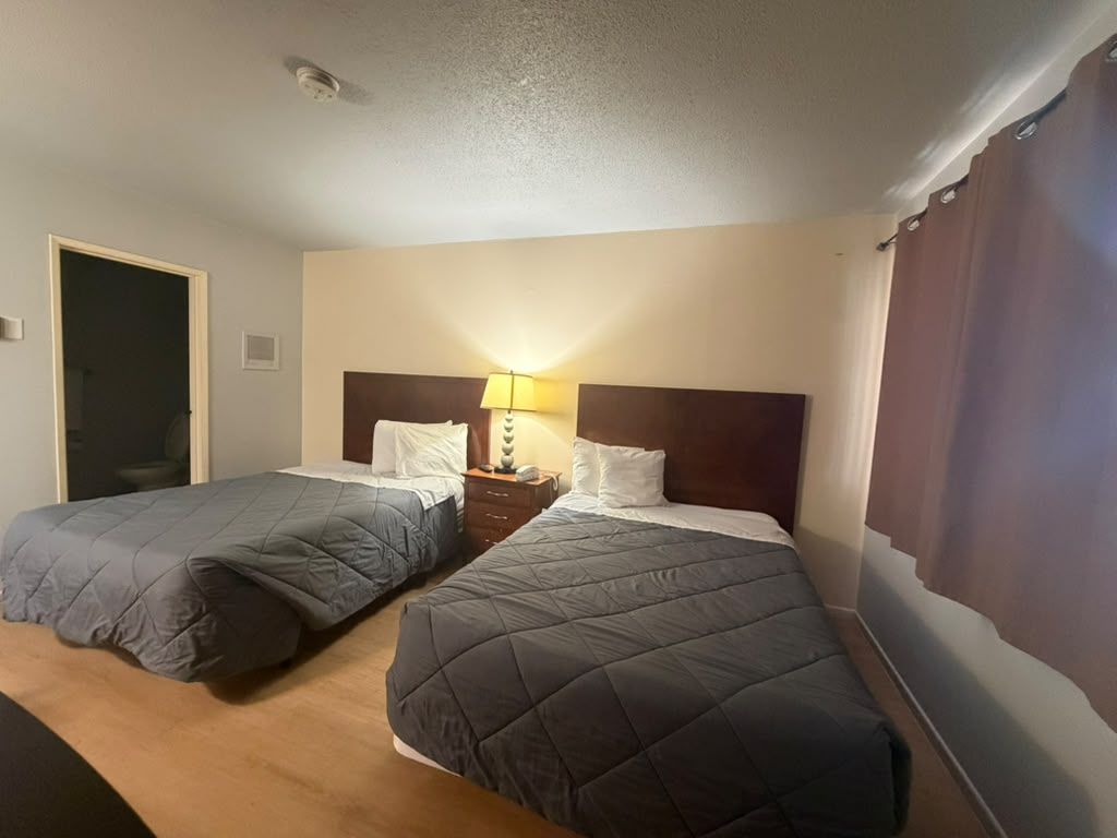 A hotel room with two beds, a nightstand with a lamp, and a door.