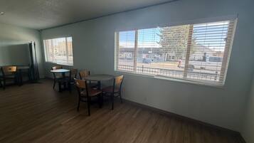 Una habitación con mesa y sillas, ventanas amplias con persianas y vista a la calle.
