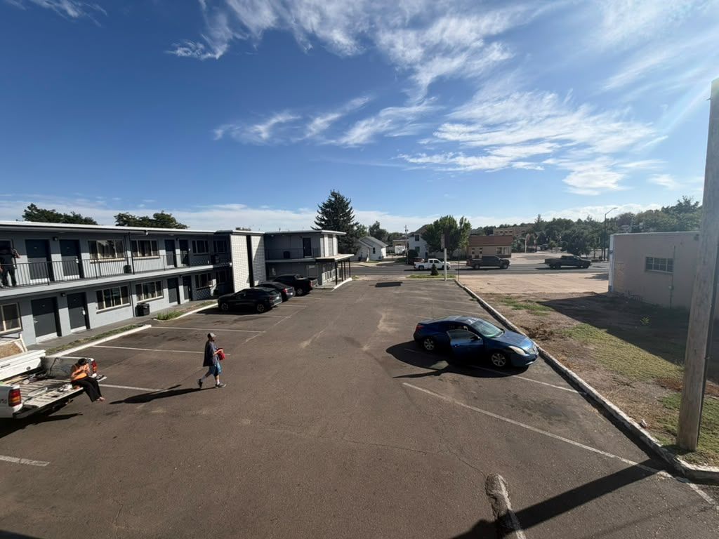 A parking lot with a building in the background, a blue car parked, and a person walking.