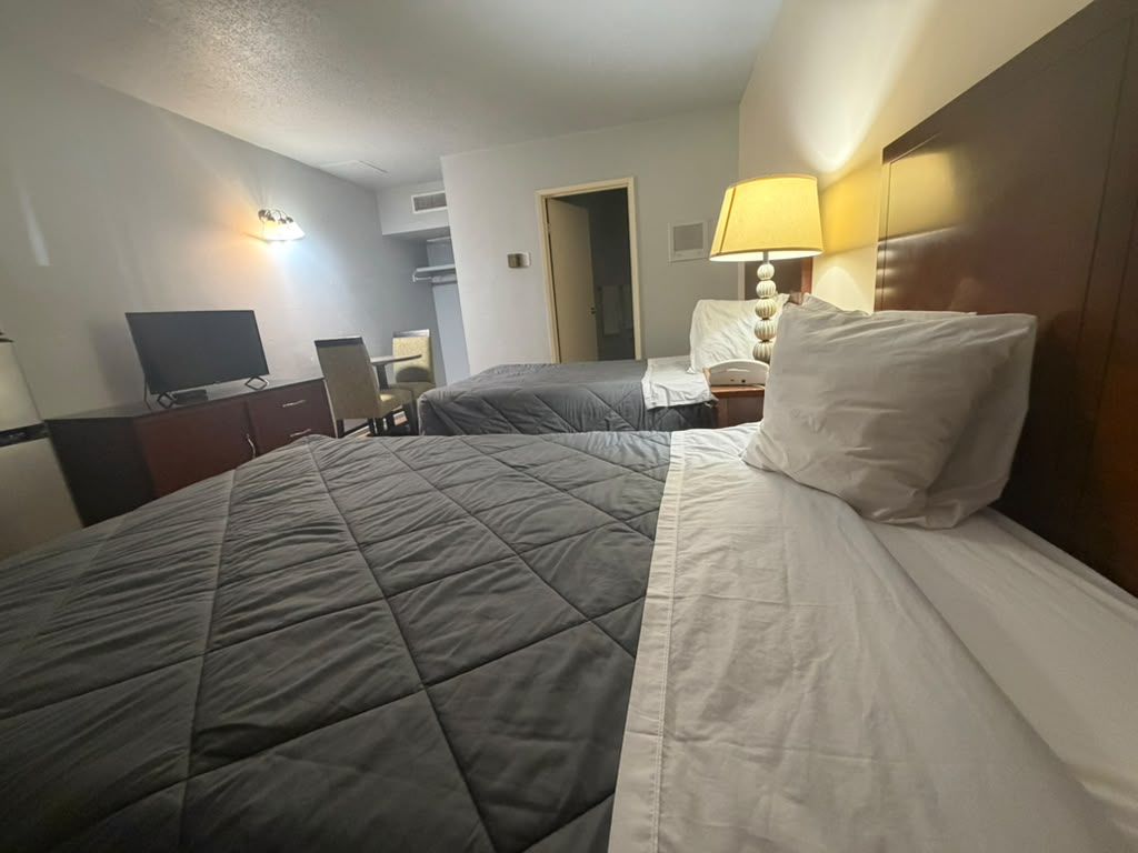 Quarto de hotel com uma cama, uma escrivaninha, uma cadeira, uma televisão e um abajur.
