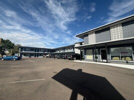 Un edificio moderno con techo plano y amplias ventanas, un estacionamiento con varios coches, y un cielo despejado.