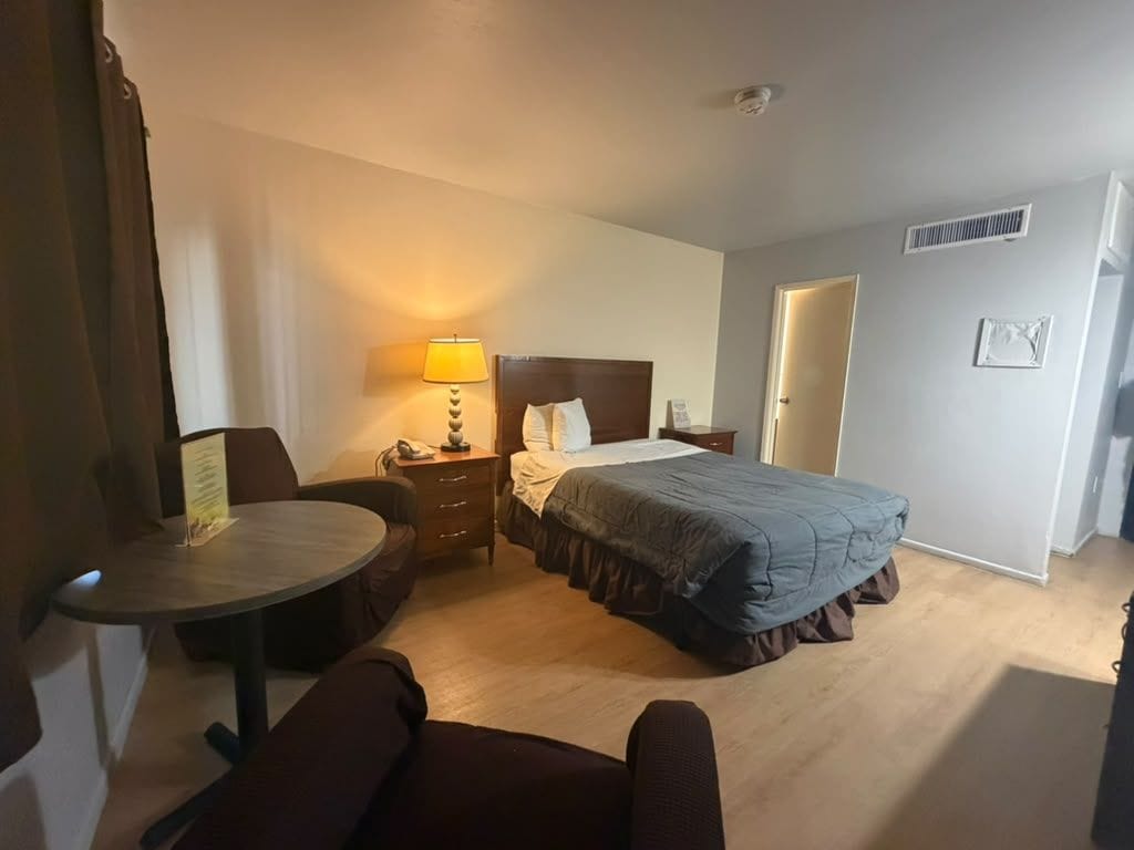 A hotel room with a bed, bedside table, lamp, and a small round table with a chair.
