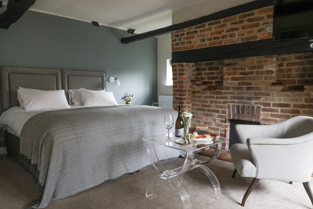A bedroom with a bed, a glass coffee table with a bottle and glass, a chair, and a brick fireplace.