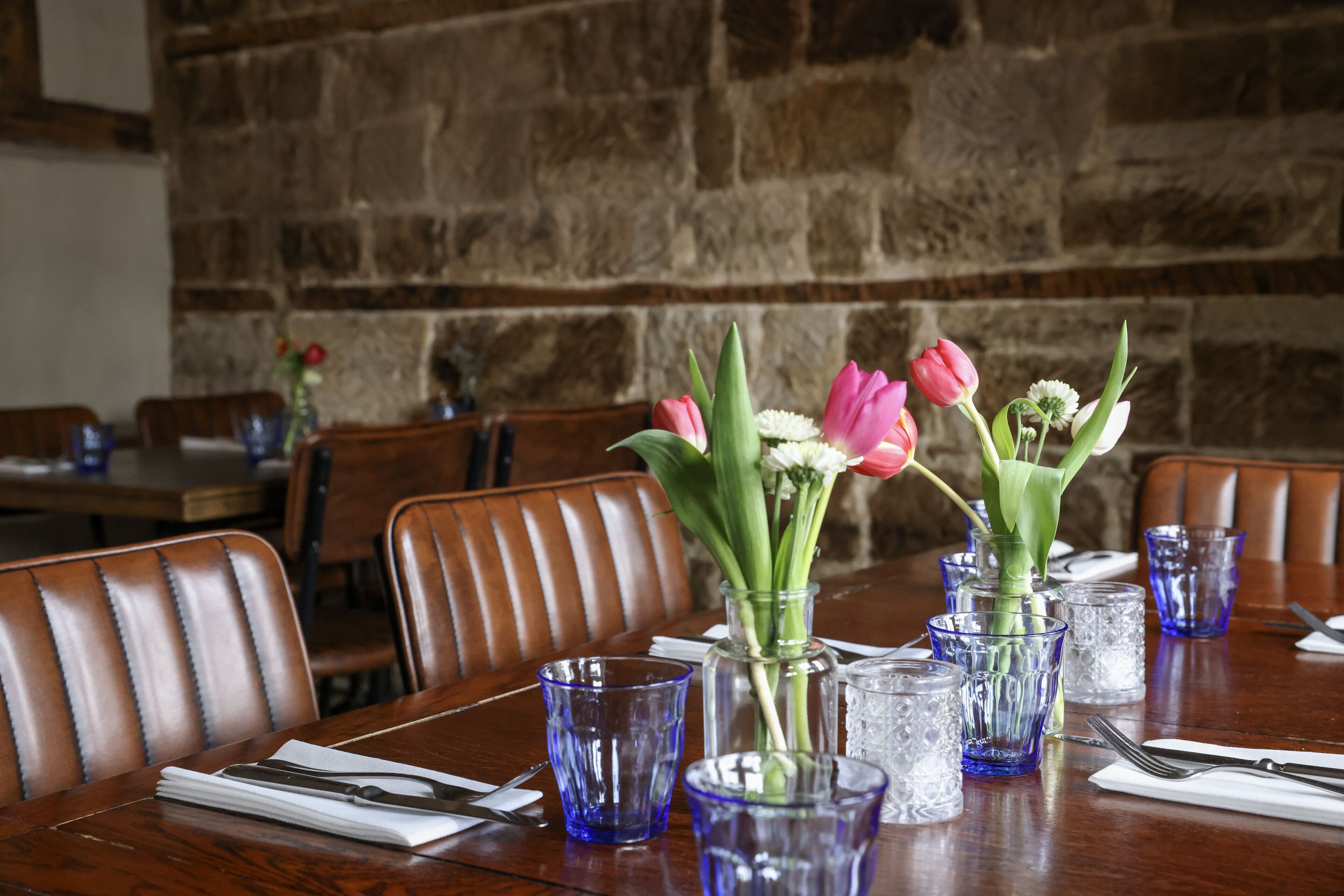 A dining table with blue glassware, a vase with flowers, and a stone wall in the background.