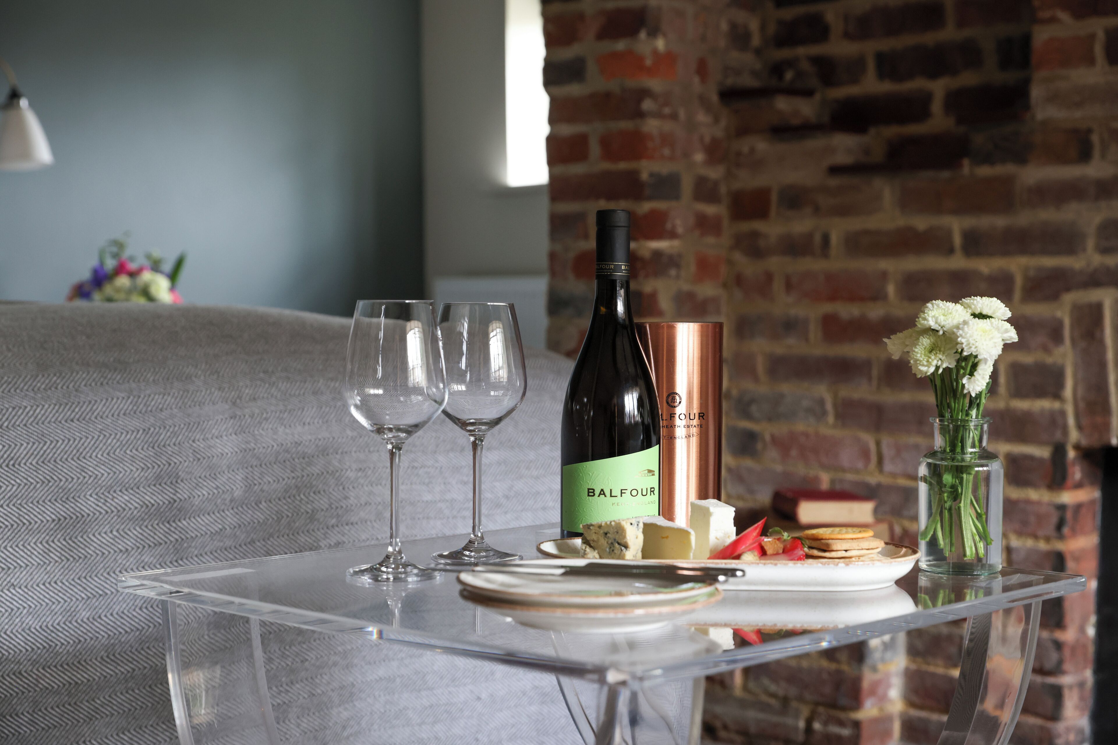 Una mesa de vidrio transparente con una botella de vino Balfour, copas, queso y un florero con flores.