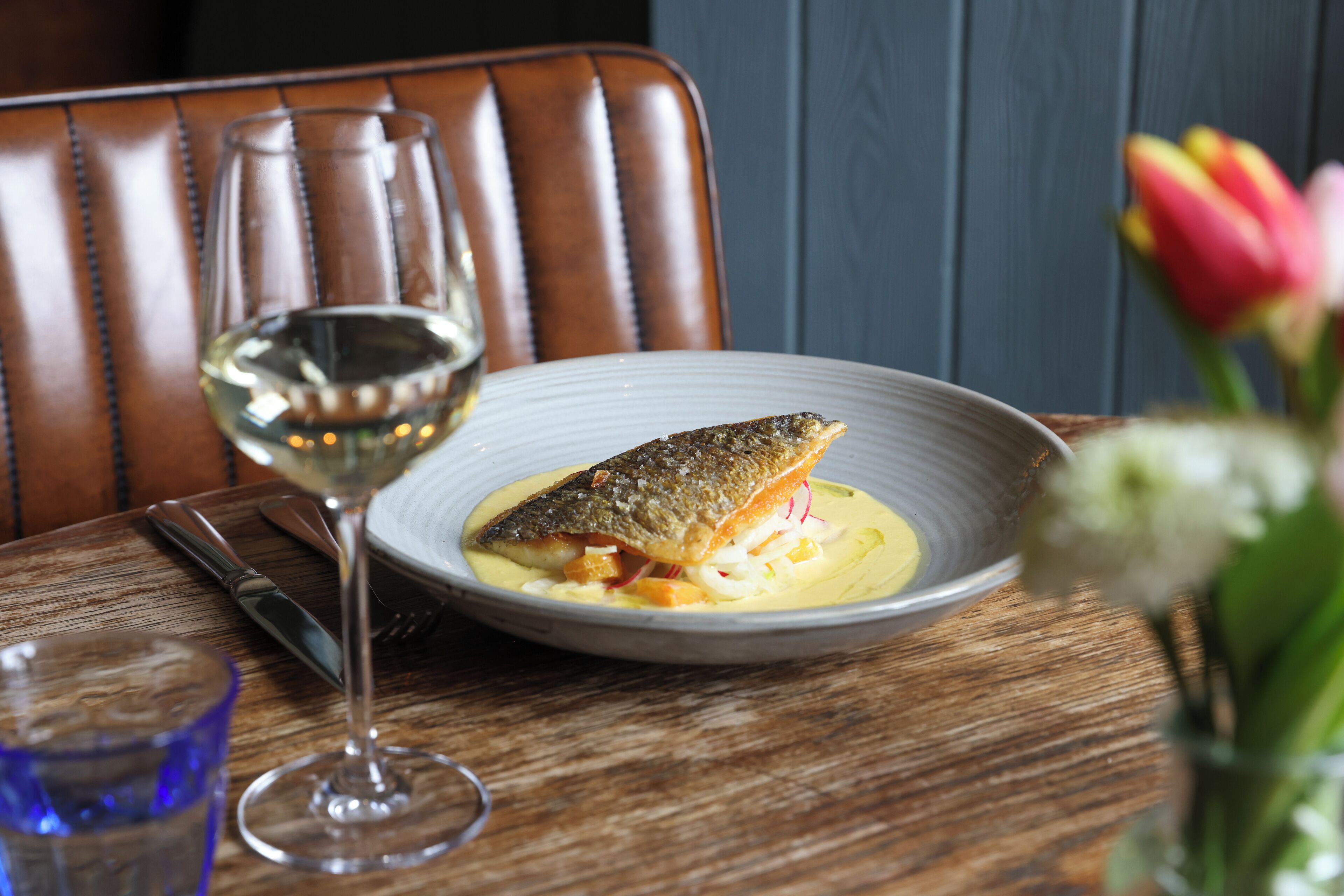 Una mesa de comedor con un plato de pescado y una copa de vino blanco.