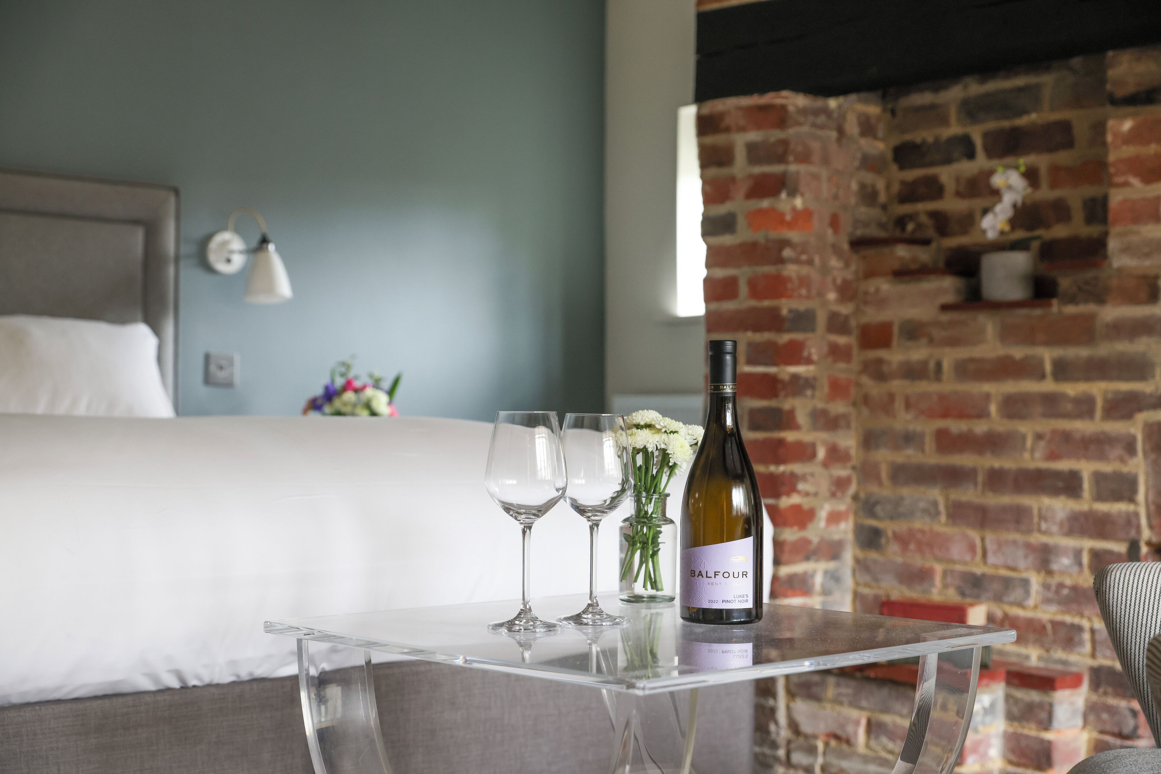Une chambre d’hôtel avec un lit, une table dégagée sur laquelle se trouvent une bouteille de vin et des verres, et un mur en briques.