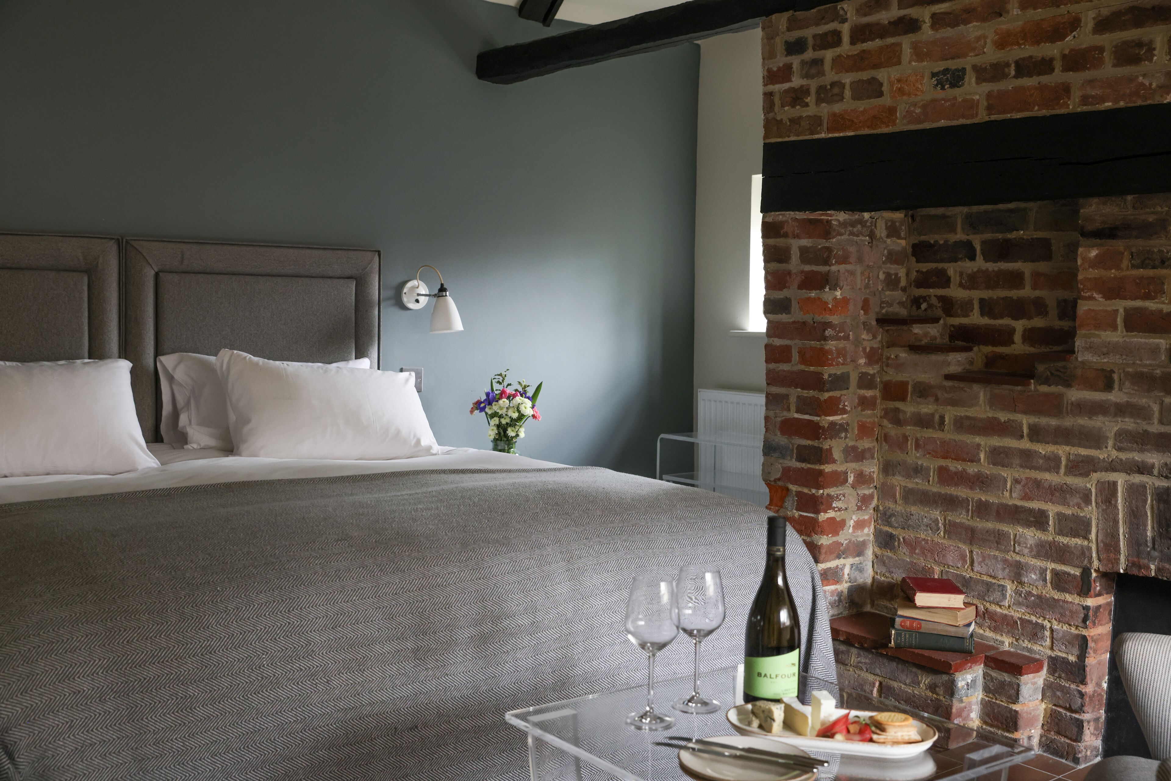 A neatly made bed with a grey bedspread, a bedside table with a bottle and glasses, and a brick fireplace.