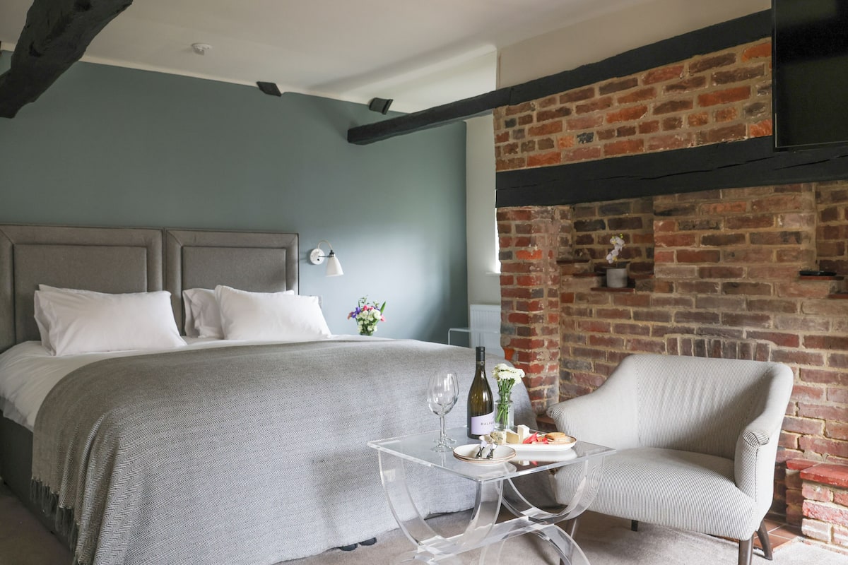 A bedroom with a bed, a chair, a table with a wine bottle and flowers, and a brick wall.