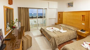 Een hotelkamer met een bed, een bureau, een stoel, een klein tafeltje met een bloevas, en uitzicht op het strand door een glazen deur.