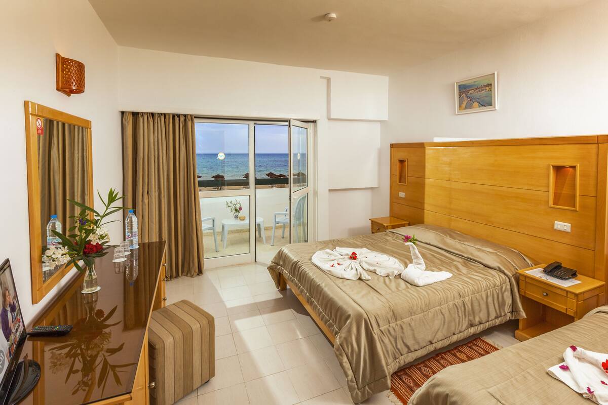 Een hotelkamer met een bed, een bureau, een stoel, een klein tafeltje met een bloevas, en uitzicht op het strand door een glazen deur.