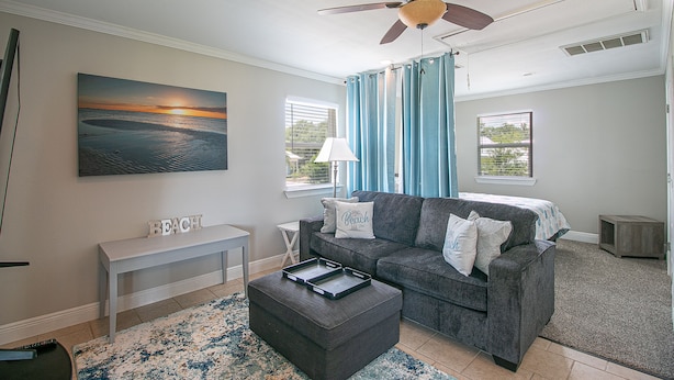 A living room with a gray sofa, a white bench, a painting of a beach, and a ceiling fan. - Flirty Flamingo, Too! 1 BDR 1BTH, with Shared in-ground pool! (Gulfport)