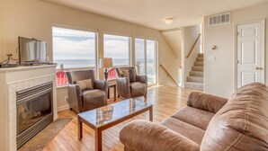 A spacious living room with a fireplace, leather sofas, a coffee table, and a television. Large windows offer a view of the ocean. - Storm Watchers Pointe (Lincoln City)