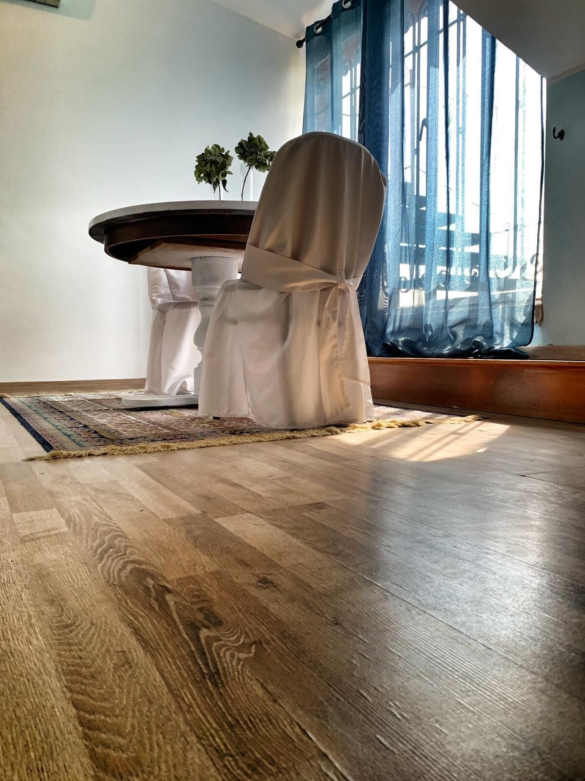 A room with a wooden floor, a white chair with a white cover, a small table, and a window with blue curtains.