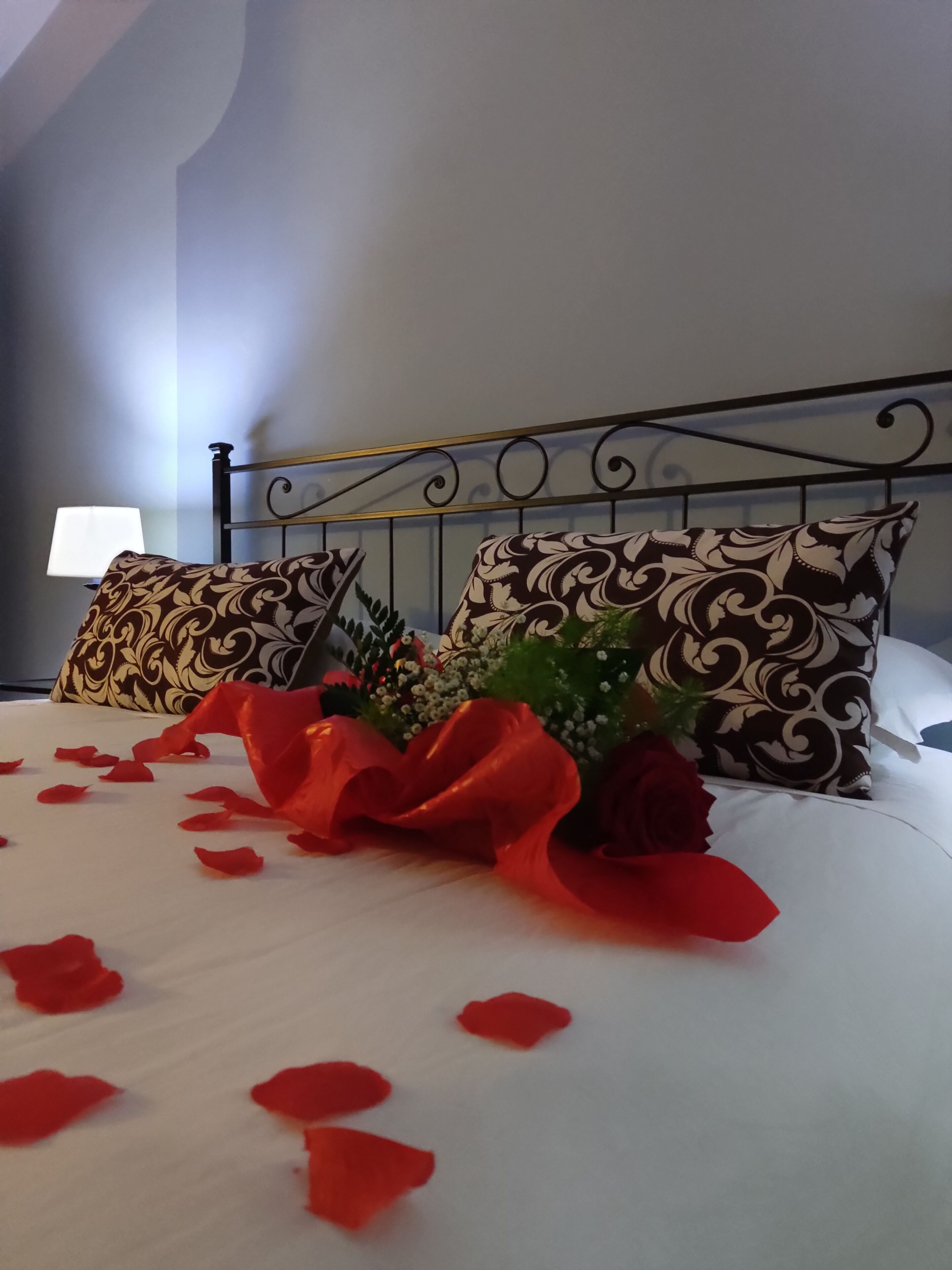 Una cama cuidadosamente hecha con pétalos de rosa y un lazo rojo, dos almohadas estampadas y una mesita de noche con una lámpara.