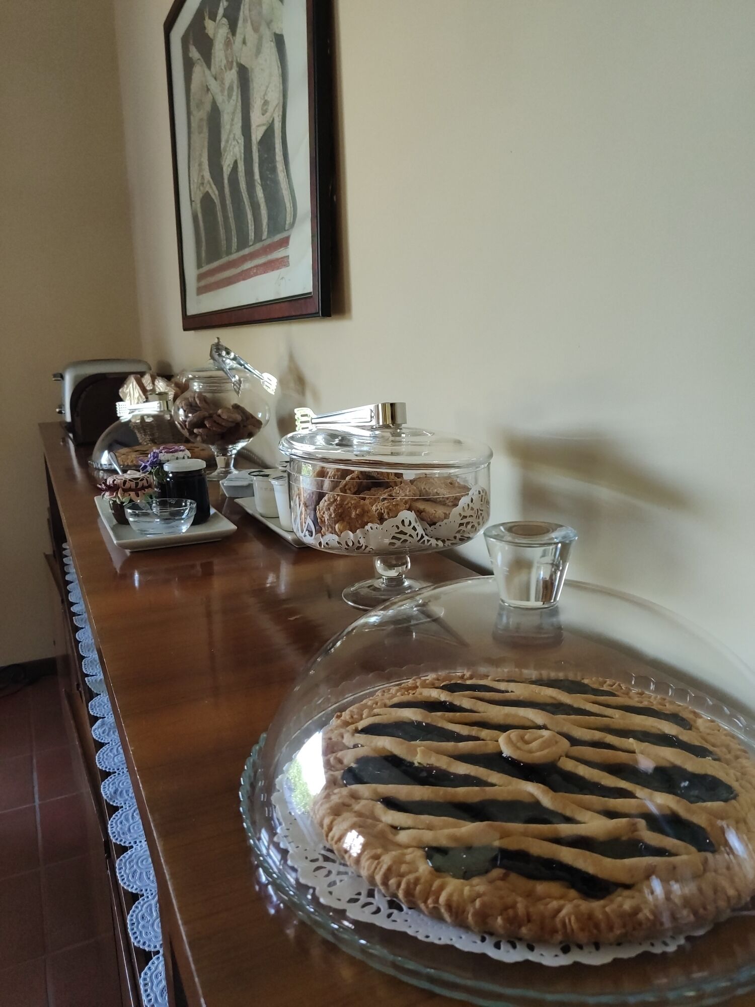 A wooden table with a glass pie on a doily, a jar of cookies, a glass of milk, and a framed picture on the wall.