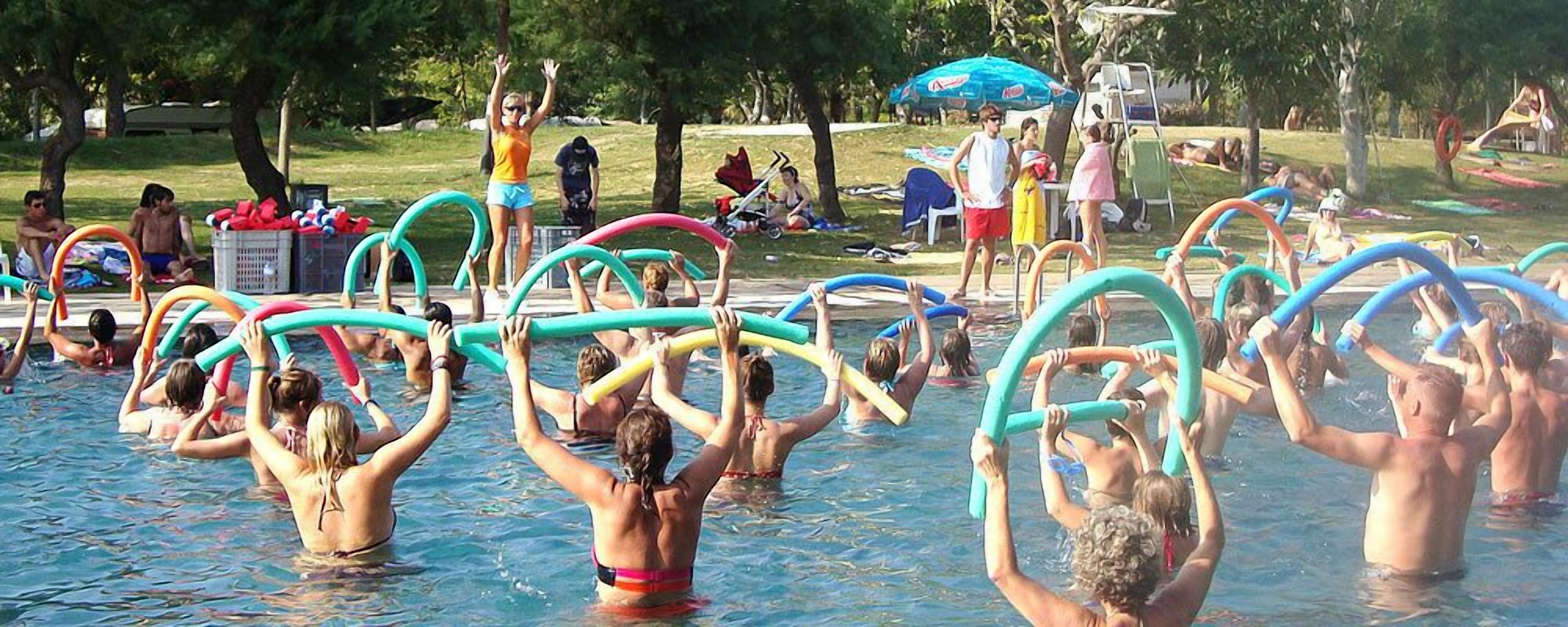A swimming pool area with people engaging in water activities, surrounded by trees and shaded by umbrellas.