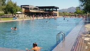 A swimming pool with people in the water, a wooden deck, and a building in the background. - Rubina Resort (Castello d'Empuries)