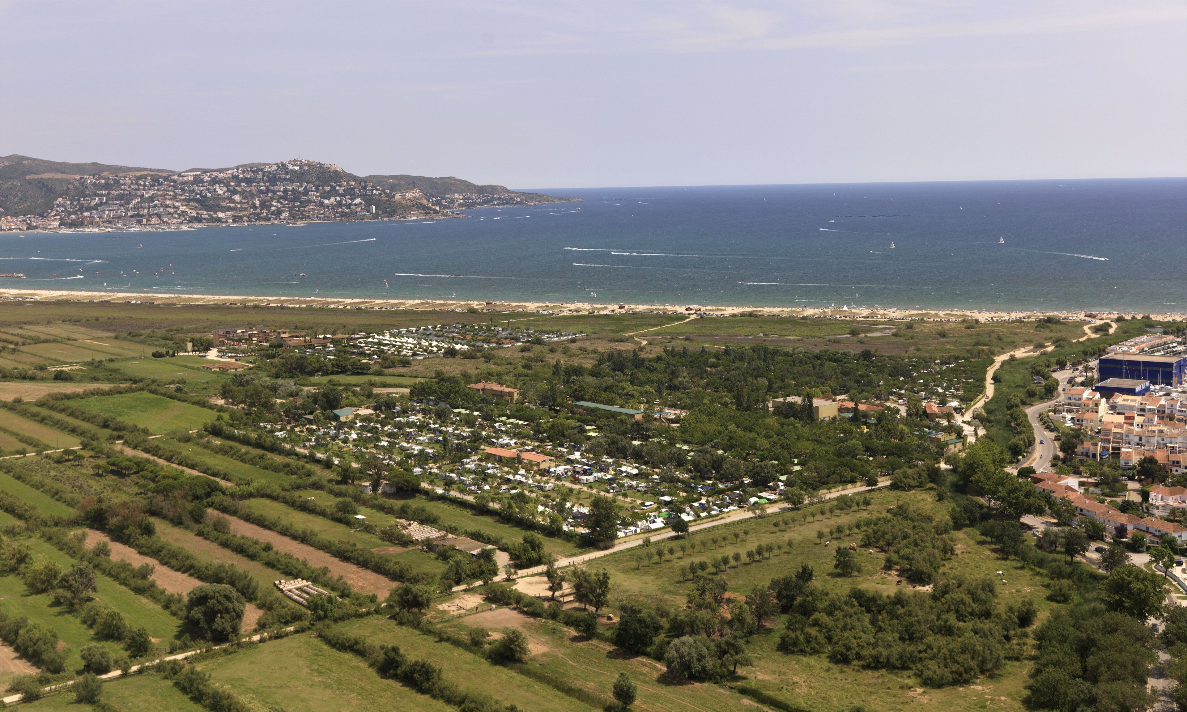 Vista aérea de uma vila costeira com praia, áreas residenciais e campos verdes.