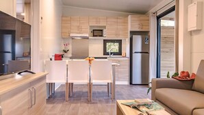 A modern kitchen with wooden cabinets, a microwave, and a refrigerator. There is a dining area with white chairs and a table. A sofa and a coffee table are visible in the living area. - Rubina Resort (Castello d'Empuries)