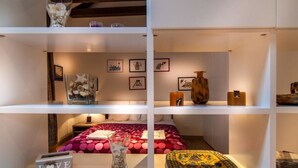 A hotel room with a bed, a shelf with decorative items, and a wall with framed pictures.