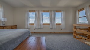 Une chambre à coucher avec un lit, une table de chevet en bois, un bureau et trois fenêtres ornées de rideaux blancs.