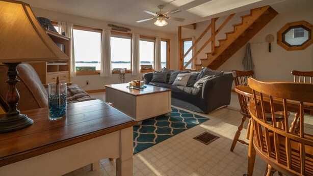 Un salon chaleureux avec une table basse en bois, un canapé et une table à manger avec des chaises. Un escalier mène à un étage supérieur, et de grandes fenêtres offrent une vue sur l’extérieur.