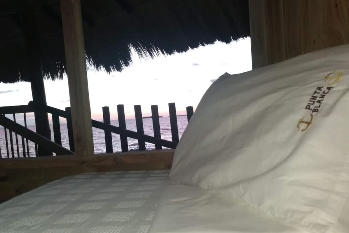 A bed with a pillow labeled "PUNTA BLANCASA" and a view of the ocean through a wooden balcony.