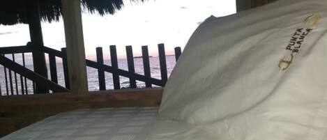 A bed with a pillow labeled "PUNTA BLANCASA" and a view of the ocean through a wooden balcony.