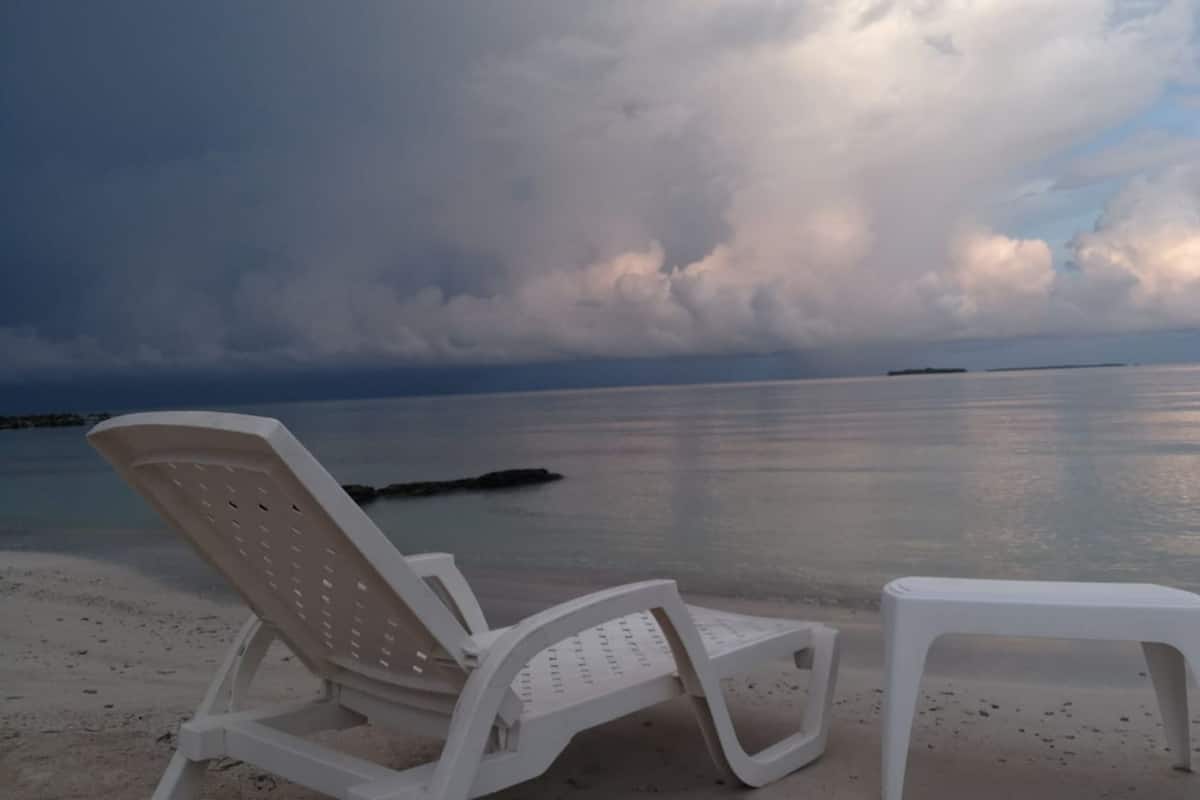 A beach with a white plastic lounge chair and a small table, overlooking calm waters and a cloudy sky.