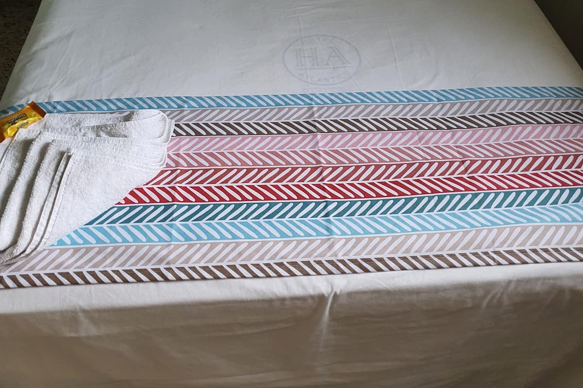 A single bed with a white mattress and a patterned blanket. There is a pillow with a striped design and a white pillow. A towel is placed on the bed.