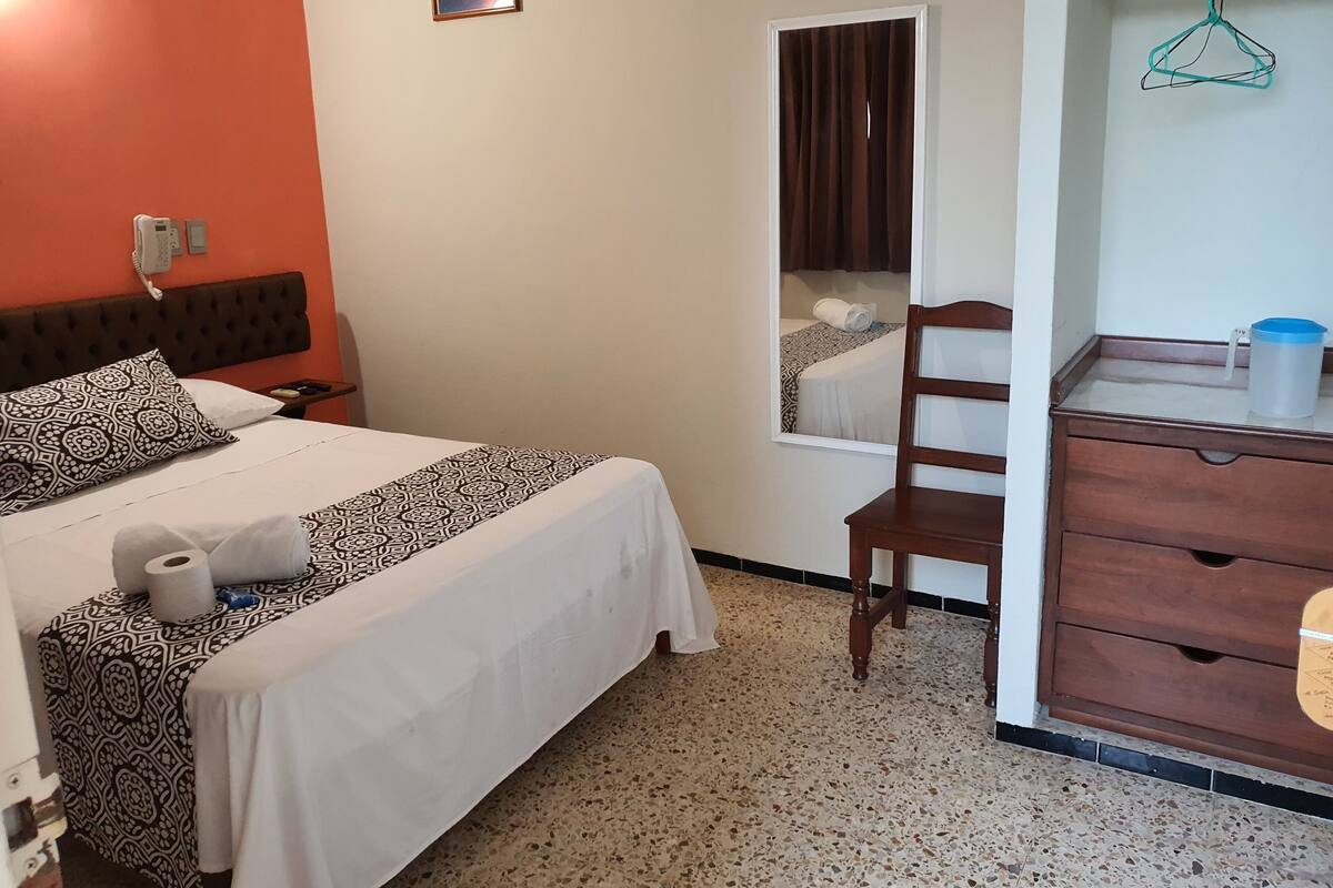 A hotel room with a bed, a wooden dresser, a chair, a window with curtains, and a small mirror.