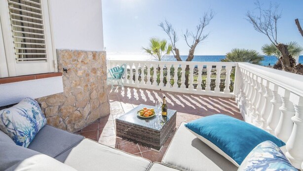 Ein Balkon mit Steinmauer, weißem Geländer und Blick aufs Meer. Ein Korbstuhl mit Flasche und Obst, sowie ein blaues Kissen auf der Couch.