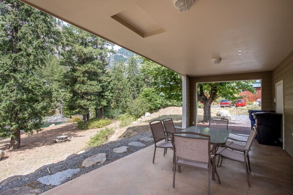 A covered patio with a table and chairs, overlooking a natural landscape with trees and a gravel path.