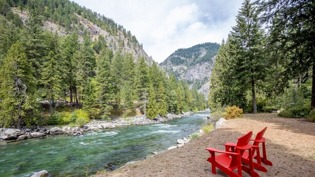 A river with a rocky bed, surrounded by dense green trees and a mountain in the background. There are two red Adirondack chairs on a dirt area.
