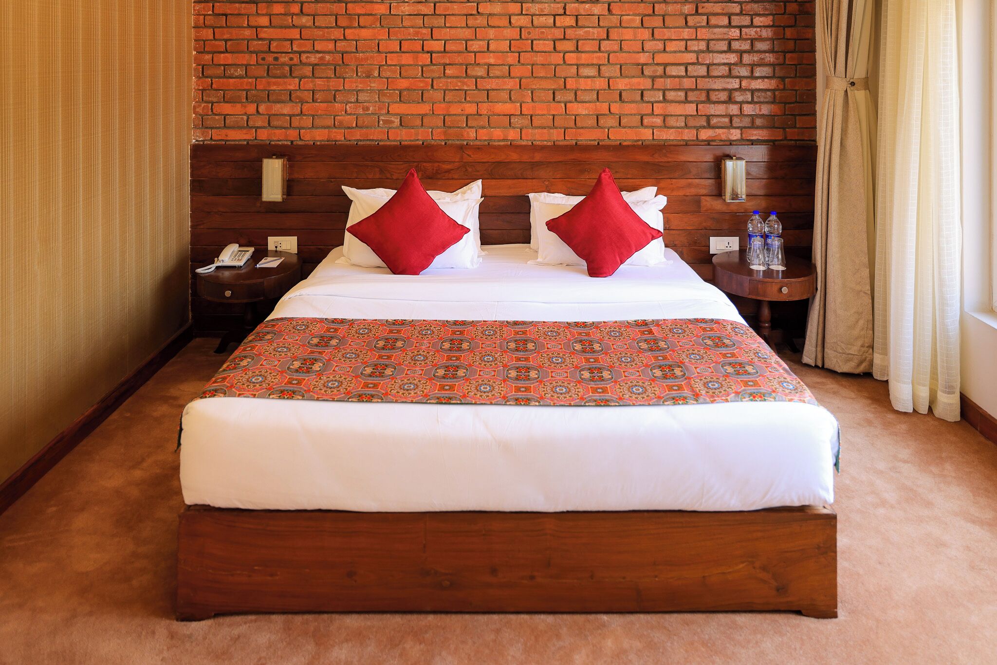 A hotel room with a large bed, a brick wall, a nightstand with a phone, and a side table with water bottles.