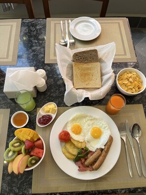 Una mesa de mármol con tostadas, cereal, huevos, tocino y fruta para el desayuno.