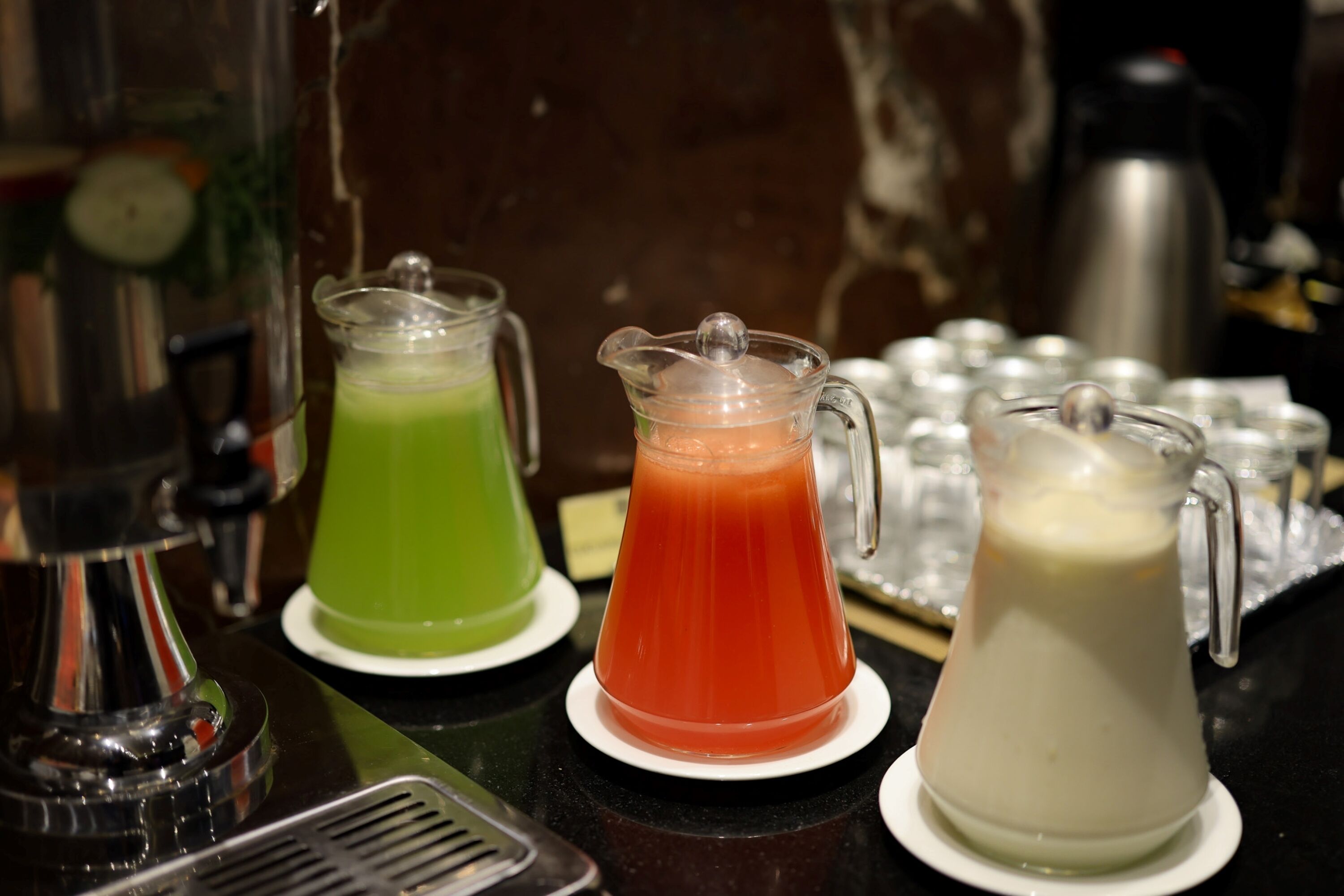 A bar area with a coffee machine, three pitchers of different colored beverages, and a metal pitcher on a counter.