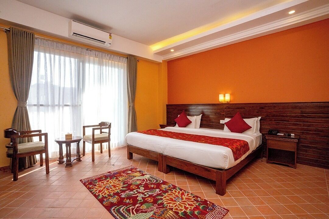 A hotel room with a large bed, two bedside tables, a small table, two chairs, a patterned rug, and a window with curtains.