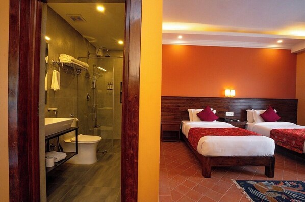 Habitación de hotel con baño que incluye cabina de ducha con mampara de vidrio, lavamanos y retrete. La habitación tiene dos camas con ropa de cama roja y cabeceros de madera.