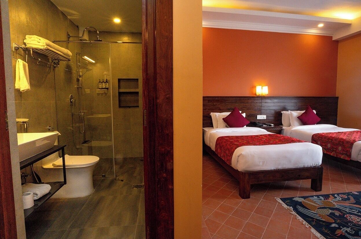 A hotel room with a glass-enclosed shower, a wooden headboard, two beds with red pillows, and a tiled floor.