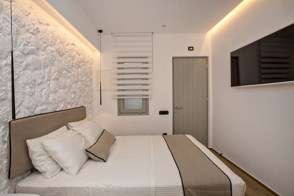 A bedroom with a bed, a headboard, a window with blinds, a door, and a wall-mounted TV.