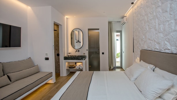 A modern bedroom with a bed, a sofa, a mirror, and a bathroom with a sink and towel rack. - Levantes Luxury Suites II (Tinos)