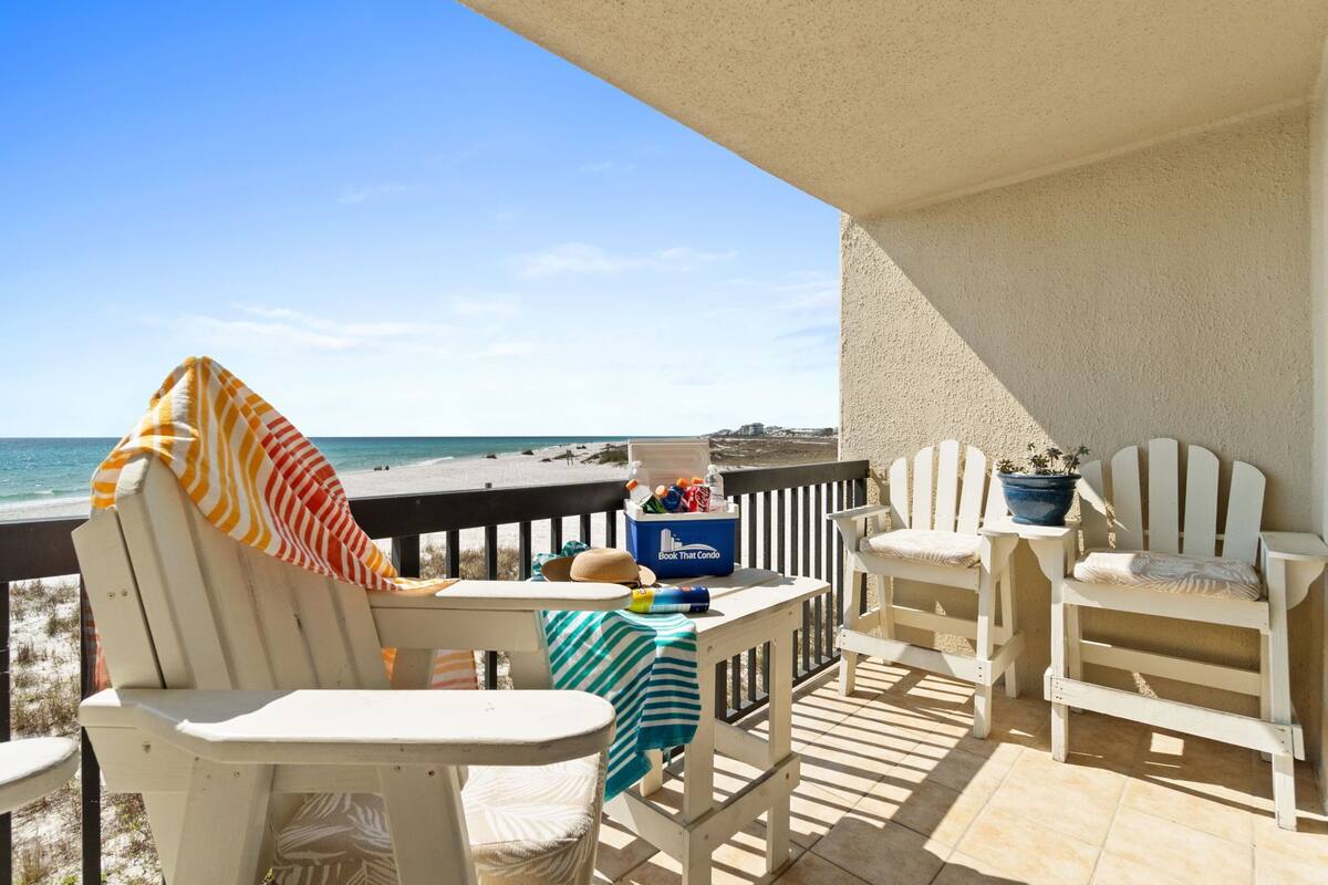 Ein Balkon mit weißen Adirondack-Stühlen, einem gestreiften Handtuch, einer Kühlbox mit Getränken und Blick auf Strand und Meer.