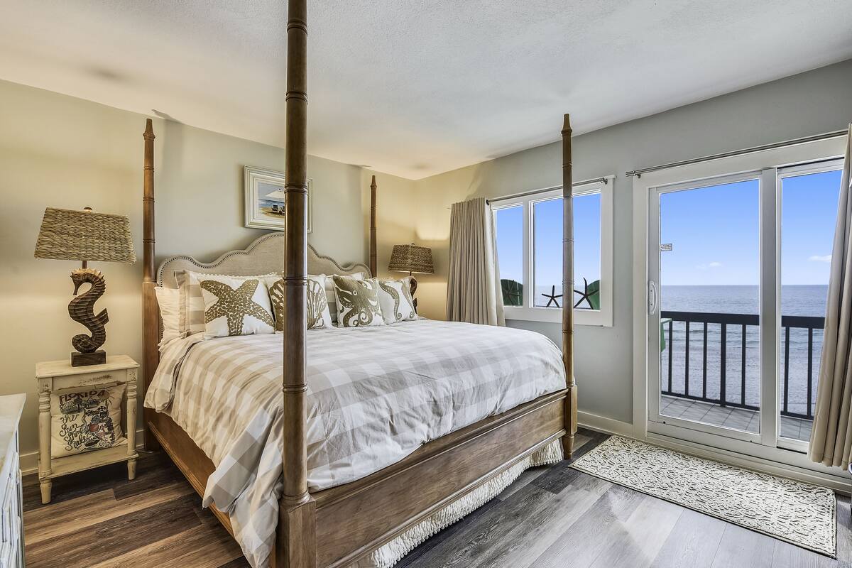 Ein Himmelbett mit kariertem Überwurf, ein Nachttisch mit Lampe und Blick auf das Meer durch große Schiebetüren.