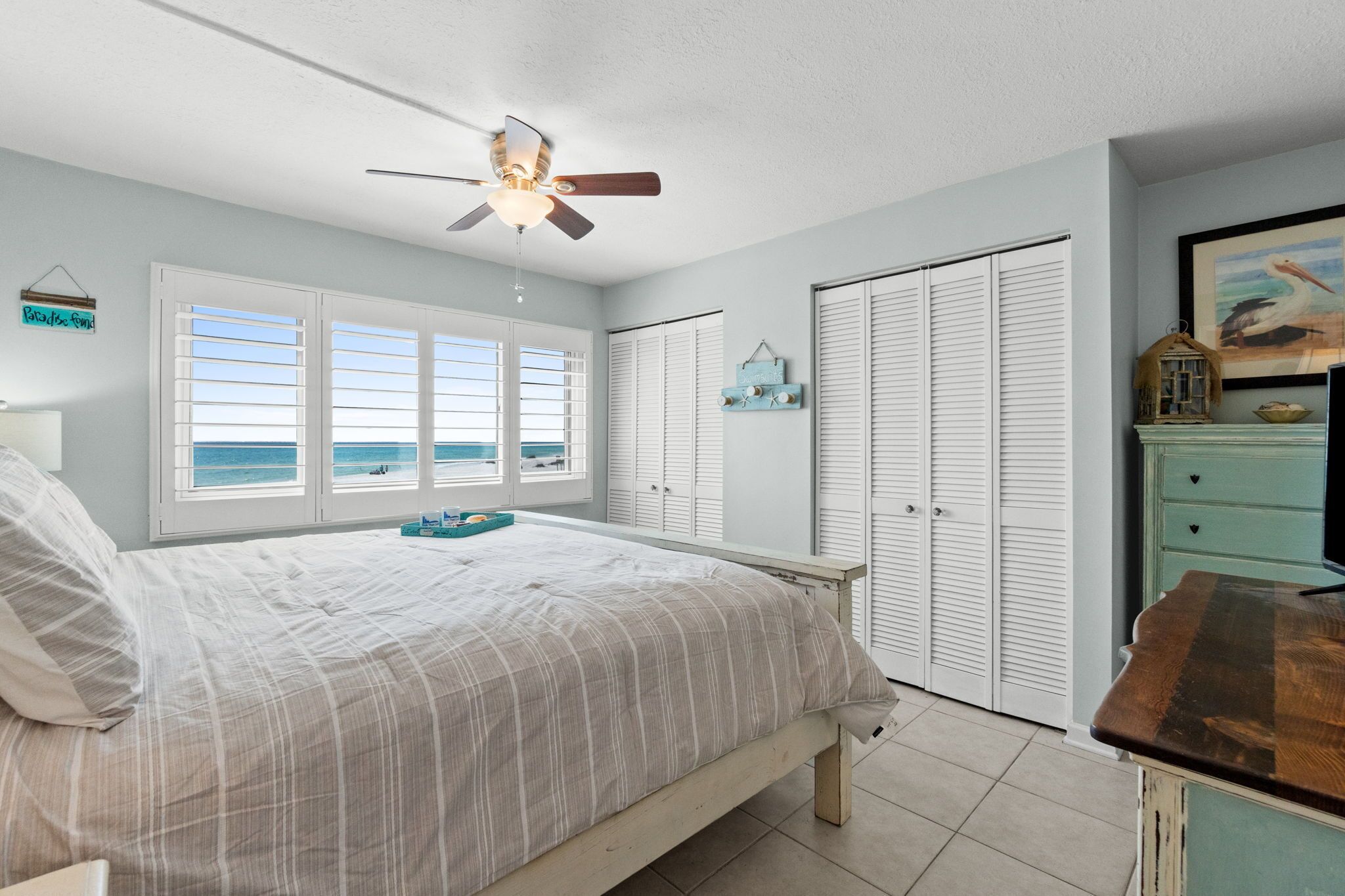 A bedroom with a bed, a ceiling fan, a view of the ocean, white shutters, and a dresser.